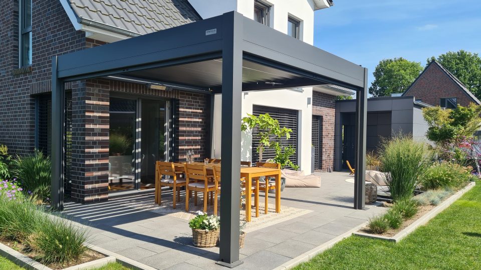 Terrasse unter einer anthrazitfarbenen Pergola mit Holztisch vor einem Klinkerhaus, daneben Gräserbeete und Rasen