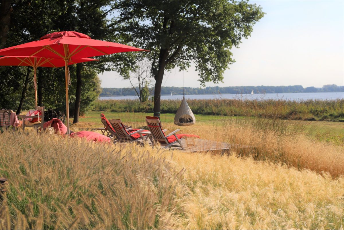 Rote Sonnenschirme und Liegestühle auf dem Holzdeck, vorn goldene Gräser, dahinter ein Hängesessel und der See